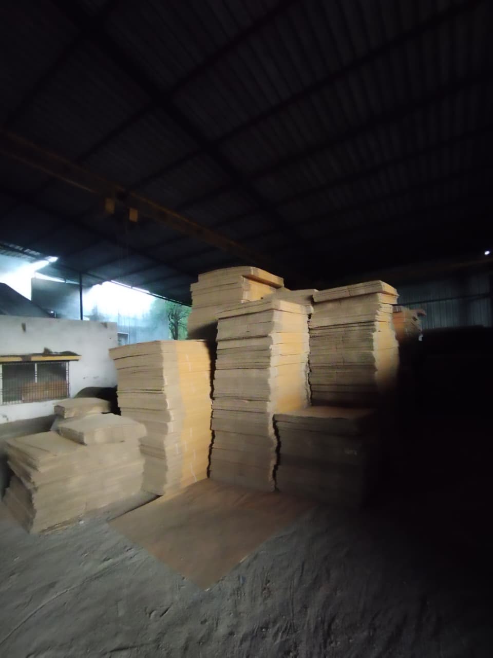 Several corrugated cardboard boxes stacked and arranged on a polished wooden floor in a bright indoor setting. Some boxes are open, showing their flaps. The environment appears clean and organized, suggesting a warehouse or packaging facility. The scene feels industrious and orderly.
